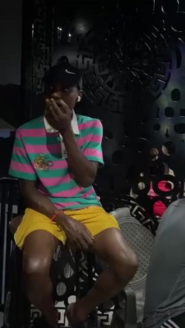 The video shows a young Black male sitting on a stool in a dimly lit room. He is wearing a pink and green striped polo shirt, yellow shorts, and a black baseball cap. He has a phone in his hand and appears to be talking. In the background, there are other people and a black decorative screen with circular cutouts. The room has a dark, modern aesthetic.