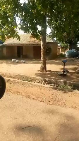 The video shows a dusty, unpaved road passing by a village. The buildings are simple, with corrugated iron roofs and some made of mud brick. In the foreground, the side mirror of a vehicle is visible. The environment appears rural and somewhat underdeveloped, with some vegetation and scattered debris. There are a few people visible in the distance, engaged in everyday activities, such as standing around or sitting. The overall impression is of a quiet, ordinary day in a village.
