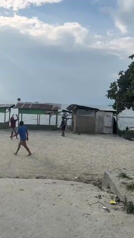 The video shows a group of children playing volleyball in a sandy area next to some small buildings. The sky is cloudy with sunlight breaking through. There are several trees in the background and a motorcycle is parked on the left side of the frame. The children are dressed in casual clothing and appear to be enjoying themselves. The scene is set in a rural or underdeveloped area.