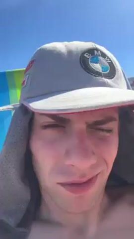 A close-up shot focuses on a young man at the beach, seated in a multicolored beach chair under a clear, bright sky. He wears a gray cap with a BMW logo and a nose ring is visible. A gray, hooded material is draped around his shoulders. His face appears slightly sunburned. Background sounds indicate music playing.
