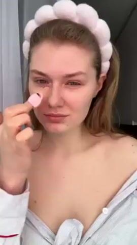 Here is a description of the video:
A young woman with reddish-blonde hair is seen applying makeup with a pink applicator in front of a window. She is wearing a light blue striped shirt with red trim and a fluffy white headband. She applies the makeup under her right eye and then on her cheek.
