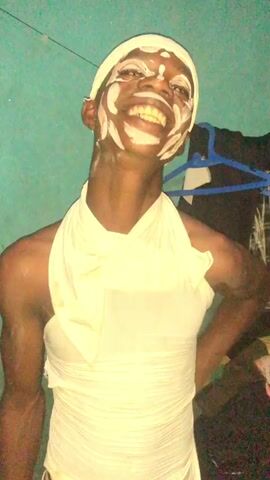 A person with dark skin and white paint on their face and body is standing in front of a turquoise wall. The person is wearing a white head wrap and a white sleeveless top that is tied at the shoulder. The person is smiling and looking at the camera. There is a clothes hanger and clothes in the background.
