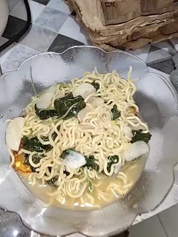 A close-up, slightly shaky, overhead shot of a bowl of instant noodles with leafy green vegetables and sliced white vegetables. The noodles are golden yellow and coiled within a clear, scalloped-edge bowl. The soup is a pale yellow color. The background is out of focus, but appears to be a countertop with a tiled pattern. To the right, a brown cardboard object is visible.