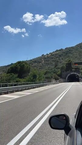 The video captures a view from a moving vehicle, looking forward along a two-lane road. The road has white double lines separating the lanes. On the left side of the road, there is a metal guardrail and a grassy embankment leading up to a hill covered in trees and vegetation. Further up the hill, there are structures that appear to be utility poles or antennas. On the right side of the road, the view is partially obstructed by the side mirror of the vehicle, reflecting a hand and arm. Ahead, the road leads into a tunnel carved into the side of the hill. The tunnel entrance is framed by stone and concrete. A red car is visible approaching the tunnel entrance from the opposite direction. Above the road and tunnel, the sky is blue with scattered white clouds. A large metal structure, possibly a traffic sign or lighting rig, is visible over the tunnel entrance. The overall impression is a scenic drive through a mountainous area.