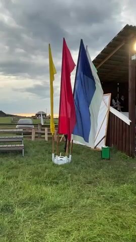 The video opens with a medium shot of an outdoor setting, panning slowly from left to right. In the foreground, three flags on poles are visible, from left to right: yellow, red, and blue with vertical white and green stripes. These flags are slightly waving in the wind. Behind the flags, a two-story wooden structure with a pitched roof stands on the right side of the frame. A bright floodlight is mounted on one of its support beams. A large, kite-like white and brown object leans against the structure, with a small green bin positioned nearby. People are seated on benches on both the lower and upper levels of the wooden structure, appearing to be spectators. Other individuals are standing and moving near the entrance to the lower level. In the background to the left, a wooden fence, additional benches, and a white car are visible, along with glimpses of distant buildings under a cloudy sky. The ground is covered in green grass.
