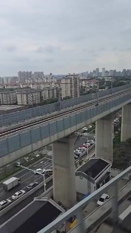 The video shows a view from a moving vehicle, likely a train, looking out over a cityscape and a raised railway line. The railway line is supported by large concrete pillars and runs across the frame. Below the railway, there is a road with parked cars and some green foliage. In the background, a dense collection of buildings forms the city skyline. The sky is overcast. The camera is moving, indicating a journey.