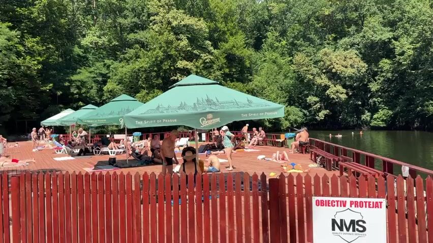 The video shows a sunny day at an outdoor lake swimming area. A wooden deck is set up along the edge of the water, with several people sunbathing and relaxing on lounge chairs and towels. Green umbrellas provide shade. A wooden fence separates the deck from the water, with a sign that reads "OBIECTIV PROTEJAT" and a logo that appears to be NMS. Several people are in the water swimming. The background is filled with lush green trees. The water is calm and reflects the trees.