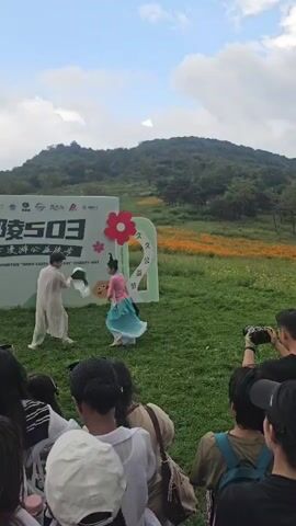 Two dancers, one in a light-colored traditional Chinese outfit and the other in a pink and teal multi-layered dress, perform with fans in a grassy outdoor area. They are positioned in front of a large white banner with Chinese text and logos, including the number "503". Behind the banner, a hill covered in trees and a field of yellow flowers are visible under a cloudy sky. A small crowd of spectators, mostly seen from the back, watches the performance, with some holding cameras.