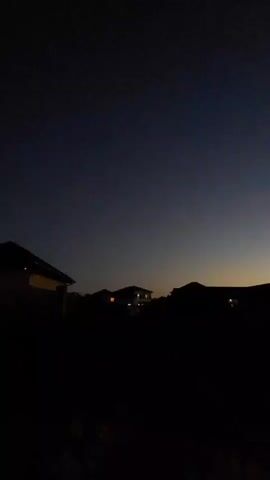 The video displays a dark sky transitioning from a deep blue to a lighter hue near the horizon. Silhouetted against this sky are the shapes of several houses and trees. Some windows in the houses are illuminated, suggesting interior lights are on. A wooden structure, possibly a fence or gate, is visible on the right side of the frame. The overall scene appears to be a residential area at dusk or dawn.
