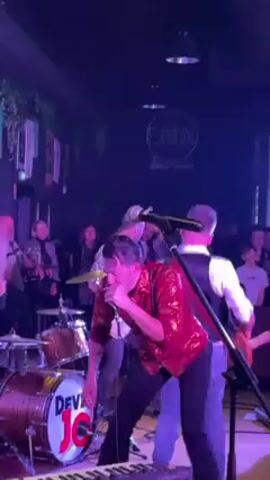 A band is performing live at a venue with an audience. The lead singer, wearing a red patterned shirt and black pants, plays the harmonica and sings into the microphone. The band includes a guitarist wearing a white shirt and vest, a drummer behind a drum kit labeled "DEVIL JO," and possibly other musicians. The lighting is dim with a spotlight on the performers. A sign for "Orbital" is visible on the back wall. The audience is watching and some are dancing.