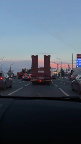 A first-person perspective video shows a view from inside a vehicle on a multi-lane road during dusk. A large red truck with two upright cargo boxes is in the center lane ahead. Several cars are visible in the lanes around the truck, all appearing to be in traffic. The sky is a gradient of blue and pink, indicating sunset. Streetlights are present on the sides of the road.