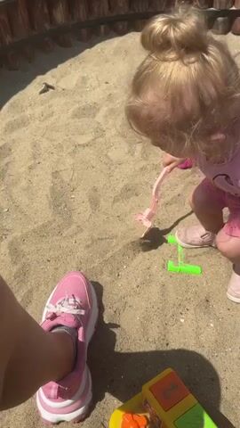 A young child with blonde hair tied in a bun, wearing a pink t-shirt and pink shorts, plays in a sandbox. The child is holding a pink toy shovel and appears to be digging in the sand. Nearby, there's a small green toy and a colorful block toy at the bottom of the frame. A person's legs, wearing pink sneakers, are visible in the foreground, suggesting they are also in the sandbox. The sandbox has a wooden border. The scene appears to be outdoors on a sunny day, with sand covering the ground.