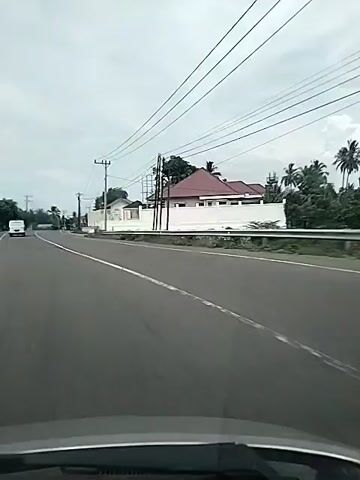 The video shows a road with a white line marking the lane. On the right side of the road, there is a white fence and some buildings. There are also power lines stretching across the sky. A white van is visible in the distance on the left side of the road. The sky is overcast.