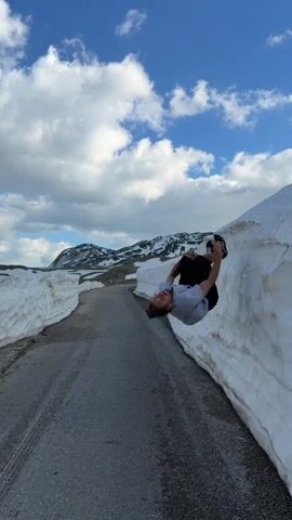 A man with a man bun is on a paved road with snow walls, seemingly having slipped and fallen, but is laughing as he pushes himself up, resting against the snow. He is wearing a grey t-shirt, dark shorts, and sneakers. The road curves into the distance with mountains visible. The sky is blue with fluffy white clouds.