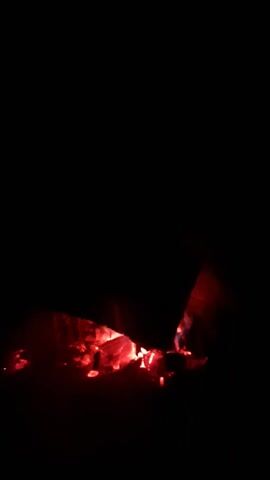 A small, contained fire burns in the dark. Red-hot embers glow brightly, emitting smoke. An object, possibly a piece of wood, partially obscures the fire.