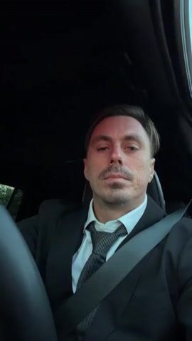 A man in a suit and tie is sitting in the driver's seat of a car, looking at the camera. The car interior is dark, with a seatbelt across the man's chest. The man has short brown hair, a mustache, and stubble. He is looking directly at the camera with a neutral expression. The lighting is dim, with some light coming from above, possibly a sunroof. There is a blurred view of green foliage in the background outside the car.