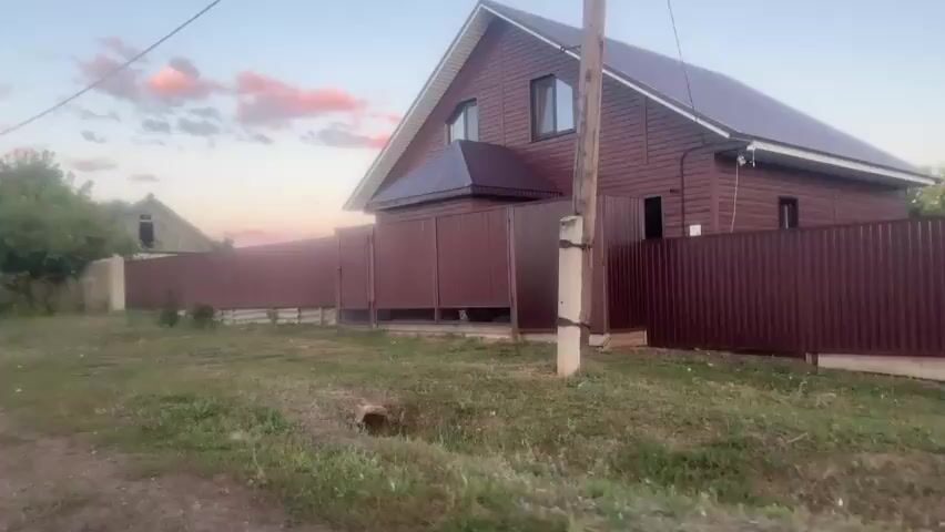 The video shows a rural scene with houses, fences, and vegetation. The camera is moving, suggesting it is from a vehicle. In the foreground, there is a dirt road and grassy areas. A large, modern house with brown siding and a dark roof is visible on the right. It is partially fenced off with a dark red metal fence. To the left of this house, there is a longer, continuous dark red metal fence that runs the length of the property. Further down the road, an older, more dilapidated house with a sloped roof and overgrown vegetation is visible behind a weathered fence. Trees and bushes are scattered throughout the scene. The sky in the background is a soft blue with some pinkish clouds, indicating it is either sunrise or sunset. A utility pole with wires is present on the right side of the frame. The overall impression is that of a quiet, perhaps sparsely populated, rural area.