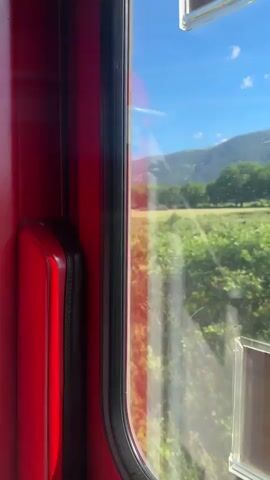 The video shows a view from inside a moving red train carriage. The camera is positioned near a window, with the red interior of the carriage visible on the left. The window reveals a bright, sunny landscape with green fields and trees. In the distance, rolling hills or mountains are visible under a clear blue sky. The train appears to be traveling through a rural or mountainous area. The footage is static with no visible people or significant actions.