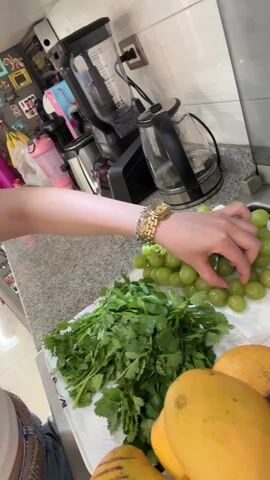 A woman in a kitchen setting holds a green grape in her hand. She brings the grape to her mouth and eats it. She then displays her hand, showing her nails painted with blue polish and white stripes. The kitchen counter holds a platter with green grapes, cilantro, and mangoes. A blender and electric kettle are in the background. A white stool is visible in the adjacent room.