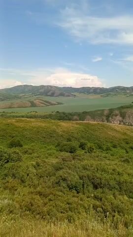 The video shows a sweeping panoramic view of a lush green landscape with rolling hills and mountains under a partially cloudy blue sky. The foreground is dominated by a dense field of green vegetation, likely grass and shrubs, with some dry grass and straw visible in the very bottom. As the camera pans to the right, the middle ground reveals more rolling hills, some with rocky outcrops, and a valley in the distance with a body of water that appears to be a river or a lake. Further in the distance, more layered mountains stretch towards the horizon. There are no people or discernible man-made objects visible in the initial frames, however as the camera pans more towards the right, a small cluster of buildings with red roofs can be spotted within the valley, surrounded by dense green trees. The overall impression is one of a serene and expansive natural environment.