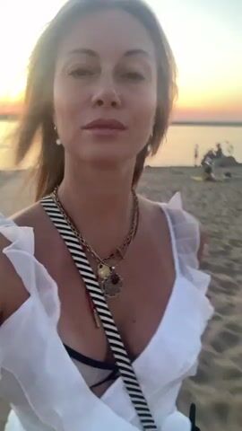 A woman with blonde hair is walking on a beach at sunset. She is wearing a white ruffled dress and a striped bag across her chest. The sun is setting over the water in the background, and there are a few other people on the beach. The woman is smiling and looking at the camera.