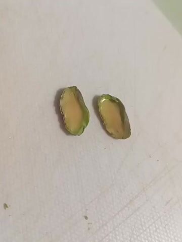 A close-up shot shows two pieces of pistachio nuts, sliced in half, on a white cutting board. The nuts have a pale yellow interior with a green outer edge.