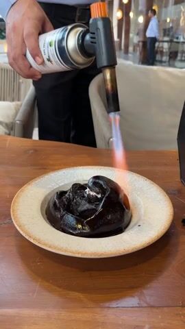 A person in a black shirt and pants is holding a culinary torch, directing the flame onto a dark, chocolate-like dessert with a dollop of white cream on top, served in a light-colored bowl. The torch is creating a visible flame and smoke as it caramelizes or sears the dessert. The dessert is placed on a wooden table. In the background, chairs and other patrons are visible in a restaurant setting.