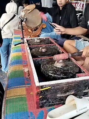The video shows three young men seated behind a gamelan instrument. The gamelan consists of three circular metal gongs set into a wooden frame, and a small drum to the side. Two of the men are seated behind the gamelan and another man is kneeling and leaning over the instrument, with his face pressed against the gongs. The third man is lying lengthwise across the gamelan with his head resting against the frame. The faces of both the kneeling man and the man lying across the gamelan are covered with dark liquid, presumably from the gongs. All three men are wearing casual clothing. People wearing civilian clothing are standing in the background. The video is shot from a close perspective.