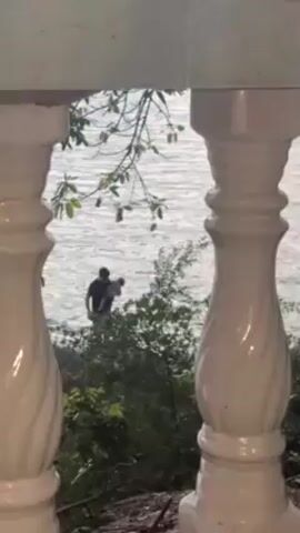 The scene is shot through the white balusters of a porch or balcony, revealing a view of a body of water. A person wearing dark clothing squats near some bushes at the water's edge, holding an object, possibly a dog. The person sets the object down on the shore.