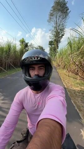 A person wearing a pink long-sleeved shirt and a black helmet rides a scooter on a paved road surrounded by tall green grass and trees. The person is looking at the camera, which is attached to the scooter. The sky is blue with some clouds. There are other people on scooters in the distance.