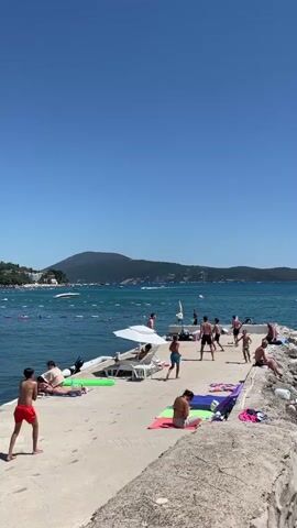 The video shows a group of people at a pier on a sunny day. The sky is clear and blue. The sea is calm, with a few boats in the distance. There are several people sunbathing and relaxing on the pier. Some people are playing in the water. A man in red shorts is walking on the pier. A woman in a white top is sitting on a towel. A man and a woman are playing with a ball. Several other people are scattered around the pier, some are sitting and some are standing. The pier is made of concrete and stone. In the background, there are hills and buildings.