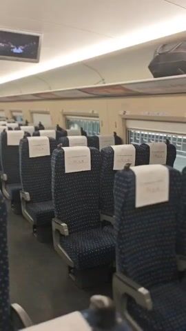 The video captures the interior of a modern train car, likely a high-speed rail, from a perspective looking down the aisle from the rear. Rows of blue, patterned seats with white headrest covers are arranged neatly on both sides of the central aisle. Overhead luggage racks are visible, with some luggage stored.

Several people can be seen in the distance, moving or standing towards the front of the train car. Two flat-screen monitors are mounted on the ceiling, displaying video content. Bright fluorescent lights illuminate the cabin. On the left side, an overhead banner or poster with red lettering is partially visible, showing Chinese characters that include "金百合" (Golden Lily) and the stock code "03678.HK," indicating a location in China. The blurred view outside the windows on the right suggests the train is in motion. The overall impression is that of a clean, well-lit, and modern transportation environment during a journey.