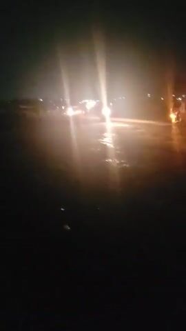 The video shows a dark scene, possibly at night. Car headlights are visible in the distance, reflecting off what appears to be a wet or glossy surface. The environment is largely undefined due to the poor lighting conditions. There are no clear indications of people or objects in the immediate foreground, only the impression of a roadside or open area.
