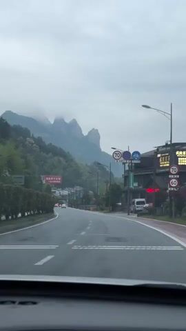 The video is shot from the interior of a car driving on a straight road. The road has white lane markings and a red bike lane on the right side. The surrounding environment is a rural area with green trees and buildings on either side of the road. In the distance, there are tall, rocky mountains partially obscured by fog. The sky is overcast. Street signs are visible along the road. Other cars can be seen driving in the distance. The windshield of the car partially obscures the view.