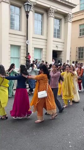 A group of people are dancing in a line formation on a street. They are holding hands and moving in a circular motion. Most of the people are women and girls, wearing colorful traditional clothing. Some of the men are also present, observing or participating. The background shows a building with classical architecture, featuring large columns and windows. The street is paved and appears to be an urban setting. The time of day seems to be daytime, with natural light.