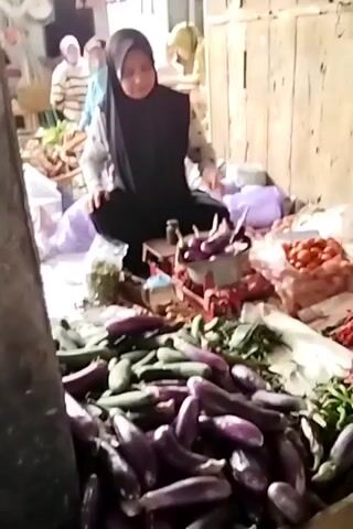 A woman wearing a black hijab and a patterned shirt kneels in a market stall surrounded by fresh produce. In front of her is a scale with eggplants on it. Piles of eggplants, cucumbers, tomatoes, and chilies are visible around her. She is holding a plastic bag in her hands and appears to be preparing it. In the background, other people can be seen at stalls selling produce. The stall itself is made of rough wood.