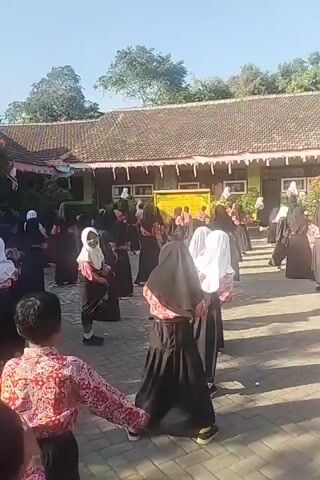 A group of people, likely students, are gathered outdoors in what appears to be a schoolyard. They are arranged in rows, facing a building with a tiled roof and a long, covered walkway. Many of the individuals are wearing dark skirts and blouses with a red and white patterned fabric, and some are wearing headscarves. They seem to be engaged in some sort of organized activity, possibly a performance or a drill, as they are all facing the same direction and appear to be in motion. The weather is sunny with a clear blue sky.