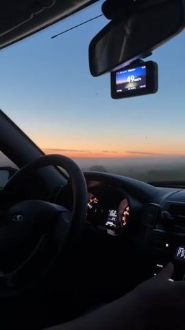 The video shows the interior of a car driving through a misty landscape at sunrise or sunset. The dashboard is visible, including the steering wheel, instrument cluster, and a dashcam displaying a speed of 48 km/h. The sky is a gradient of blue and orange. The road ahead is obscured by a thick layer of fog or mist. A triangular road sign can be seen in the distance. A hand is resting on the car's audio system.