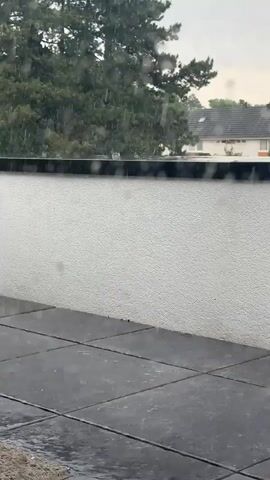 The video shows a rainy day on a balcony. Heavy rain is falling on the tiled floor and splattering against a white wall. In the background, trees and houses can be seen through the rain. As the video zooms out, a red broom and a black wicker chair come into view on the balcony. The rain is also visible on the window, creating a blurred effect on the outdoor scene.