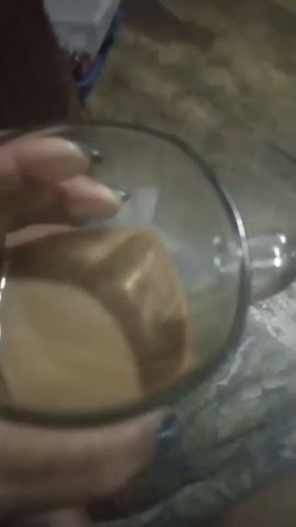 A person's hand with dark nail polish is holding a clear glass mug filled with a light brown liquid, possibly coffee or tea. The mug has a handle visible. The background is blurry and appears to be a wooden surface with some fabric or carpet. The person's bare feet are also visible in the lower part of the frame. The lighting is dim.