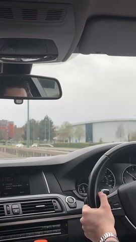 The video shows the interior of a moving car from the driver's point of view. The driver, visible in the rearview mirror, is holding the steering wheel with their right hand. The dashboard shows the car's infotainment system displaying music information: "Migos - Culture II, Walk It Talk It (feat. Drake)". The car is moving on a road with other vehicles present. In the background, buildings, trees, and road signs are visible. The weather appears to be overcast. The car seems to be approaching a roundabout. The driver is wearing a wristwatch.