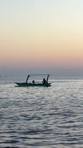 A small, green boat with a canopy is on the water. A person is sitting in the boat. The sky is a gradient of pink and yellow. There are other boats in the distance. The water is rippling.