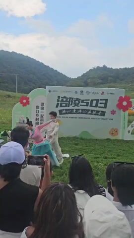 A group of people are watching a performance outdoors. In the background, there is a large banner with Chinese text and cartoon graphics. The banner appears to be for a charity event. Two performers are on a grassy stage in front of the banner. One performer is dressed in a pink outfit and the other in a light blue outfit. They are interacting with each other, and the person in blue is performing a gymnastic move, flipping over. The audience is seated on the grass, many of them holding up their phones to record the event. The environment is a grassy field with rolling hills in the background. The sky is partly cloudy.