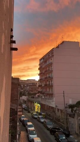 This is a vertical video clip taken from a high vantage point, looking down a narrow city street lined with buildings. The scene is bathed in the warm, vibrant hues of a sunset. The sky is a dramatic display of orange and pink clouds, creating a striking contrast with the dusky blue sky above.

The street is filled with parked cars, mostly small, compact models. A few streetlights are illuminated, casting a yellow glow on the asphalt and the surrounding architecture. The buildings are a mix of styles and ages, with some appearing older and more weathered, while others, like the prominent tall, light-pink apartment building on the right, are more modern. This building features balconies with railings and visible air conditioning units.

The overall setting appears to be an urban environment, likely residential, with a dense feel. The time of day is clearly sunset, indicated by the intense colors in the sky and the artificial lights beginning to turn on. There are no people prominently featured in the shot, but the presence of cars and buildings suggests a populated area. The emotion conveyed by the image is one of beauty and tranquility, amplified by the dramatic natural phenomenon of a colorful sunset over a city street. There is no visible text in the clip. The location is identified as Catania, Italy.