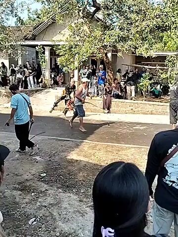 A group of people are gathered outdoors in what appears to be a village or rural setting, with a house and trees visible in the background. In the foreground, two men are engaged in a ritualistic or martial arts-like activity. One man, wearing a blue shirt, stands observing. The central action involves two men facing each other. One man, wearing a patterned garment, holds a whip-like object and appears to be in motion. The other man, with lighter-colored hair and wearing a white head covering, is also in motion, holding a similar whip-like object. Several other individuals are standing around, observing the event. The ground is a mix of dirt and asphalt. The lighting suggests it is daytime.