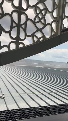 The video shows a view from inside a building, looking out at a body of water with a few boats. In the foreground, there is a ribbed metal roof or walkway. Above the water, there is a cloudy sky with patches of blue. The video pans slightly to the right, revealing more of the water and sky. The camera appears to be at an elevated position, offering a wide panoramic view. The overall scene suggests a coastal or island location.