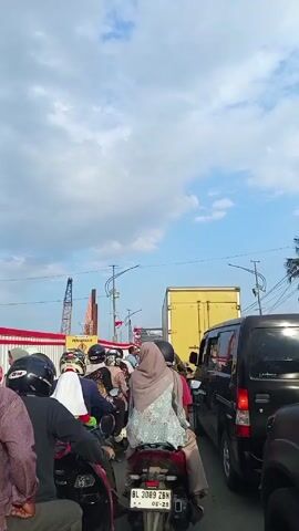 The video shows a traffic jam on a road, with various motorcycles, cars, and a large yellow truck visible. Many people on motorcycles are wearing helmets, and one individual has a hijab. A construction site is apparent on the side of the road, indicated by a red and white barrier and a construction crane in the background. The sky is partially cloudy, with patches of blue visible. The camera is positioned behind the traffic, offering a view of the road ahead. The license plate of the motorcycle in front is visible.