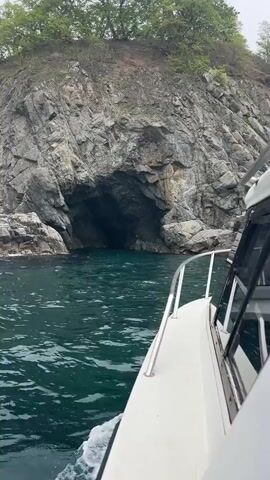 The video shows a boat approaching a large rock formation. The rock formation has a cave opening near the water level. The water is greenish-blue and appears calm.  The top of the rock formation is covered with green trees. The sky is overcast. The video is shot from the side of the boat, showing the railing and a window of the boat.