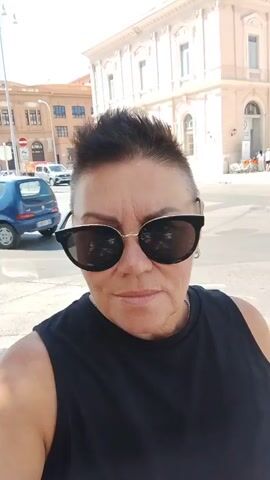 This video is a selfie-style recording of a woman with short, dark, spiked hair and sunglasses. She is wearing a black sleeveless top. The background changes throughout the video, suggesting she is moving. In the first few seconds, a building with multiple windows and a white van are visible. Later, a crosswalk and a McDonald's sign appear. Trees and a street lamp are seen in the background at one point, and in the final seconds, a truck, a scooter, and a tower-like building are visible. The lighting suggests it is daytime and sunny.