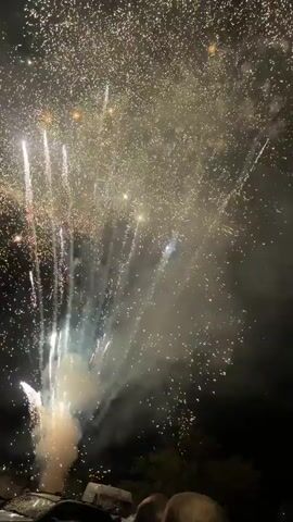 The video captures a fireworks display at night. The frame is filled with bursts of light and smoke from the fireworks. Various colors are visible, including white, gold, red, and green. The fireworks are exploding at different heights and intensities, creating a dynamic and lively scene. Some fireworks appear as sprays of sparks, while others are large, round bursts. Smoke billows in between the explosions, adding to the atmospheric effect. The overall environment is dark, indicating nighttime. In the lower part of the frame, there are glimpses of what appears to be a structure or objects, possibly the viewing area for the fireworks, but they are out of focus and indistinct. There are no people clearly visible, but the context suggests a gathering to watch the display.